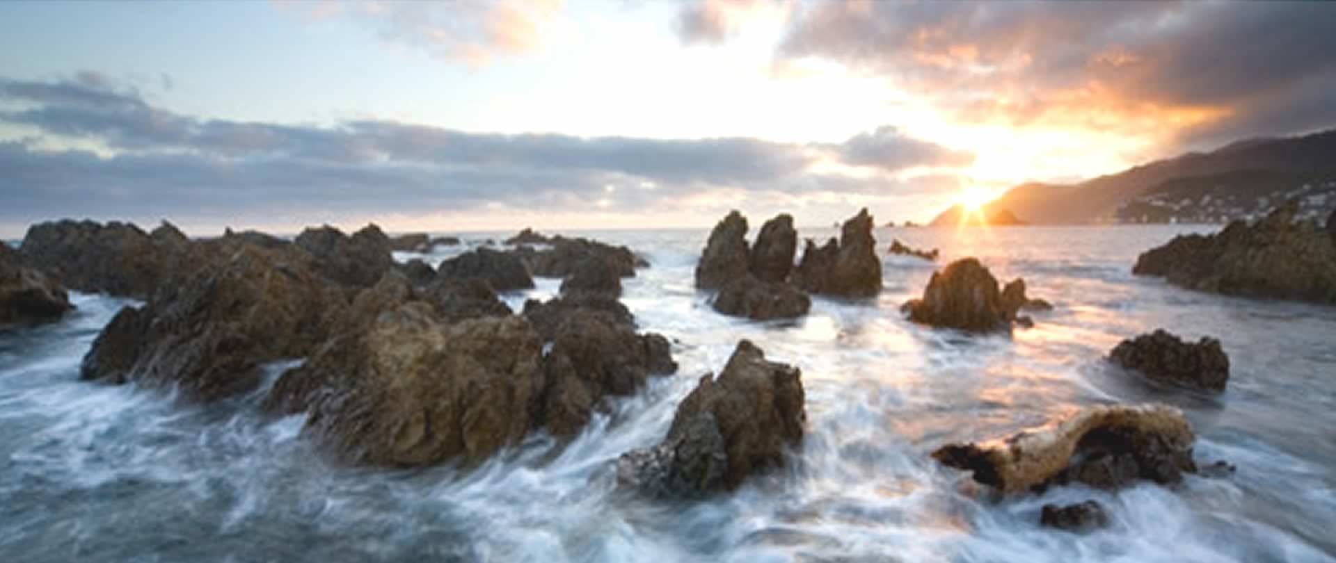 Sunset over a rocky coastline with waves crashing against the rocks.